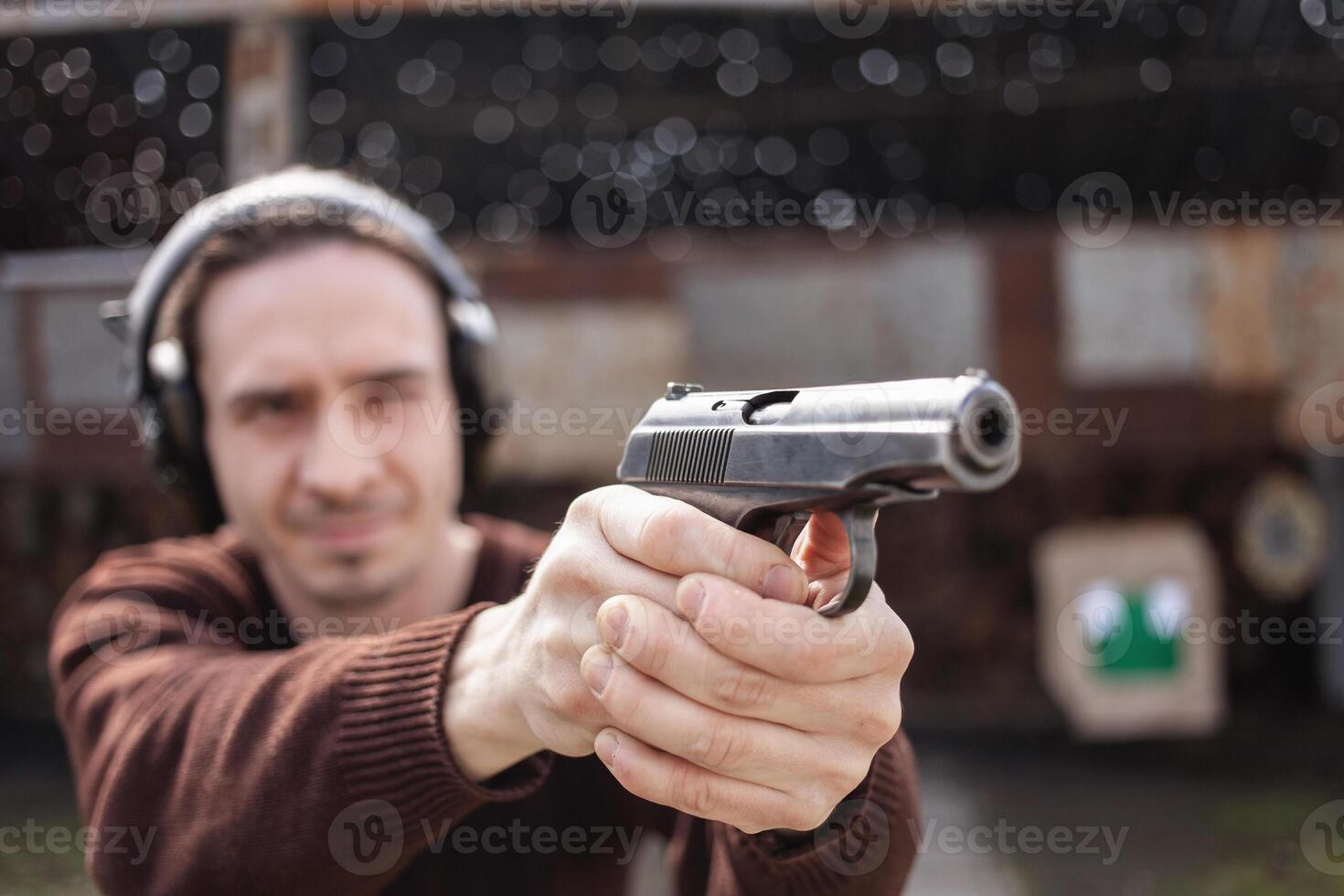 A man shoots a gun, aiming at the target. A man wearing protective headphones. A wall and a roof with bullet holes. outdoor shooting range photo