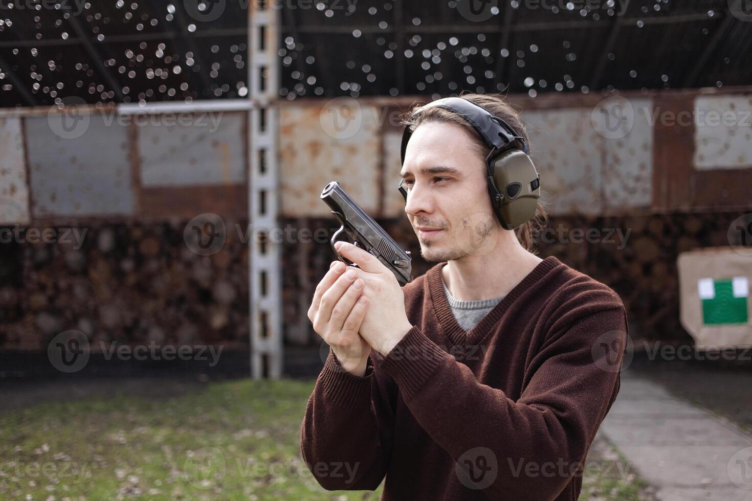 A man shoots a gun, aiming at the target. A man wearing protective headphones. A wall and a roof with bullet holes. outdoor shooting range photo