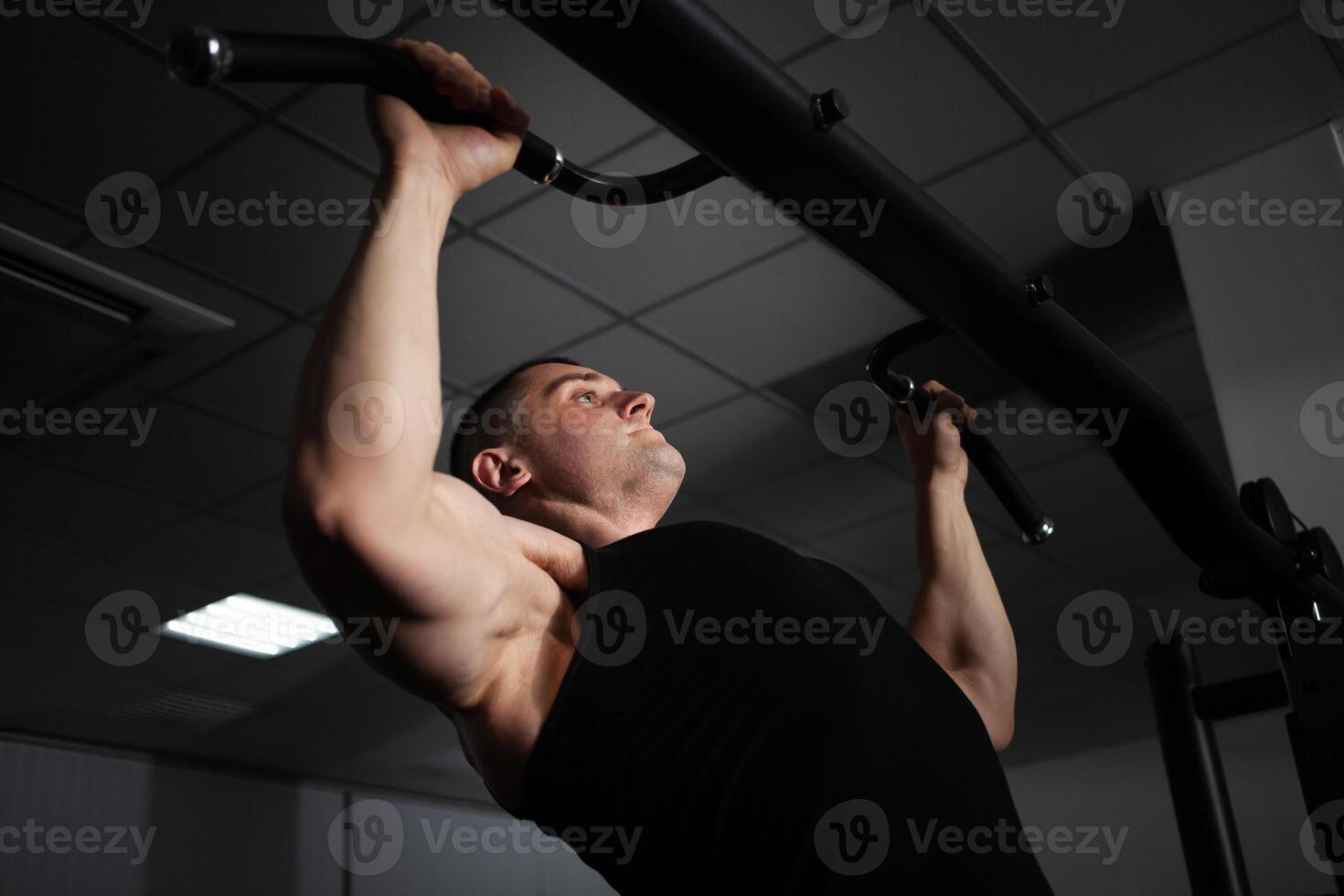 A sports athlete coach a muscular man does an exercise on a simulator in gym. Portrait, low key, darkness. pulls up photo