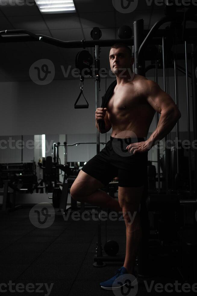 Portrait of a beautiful male athlete bodybuilder in gym, low key, darkness. Posing, coach. standing photo