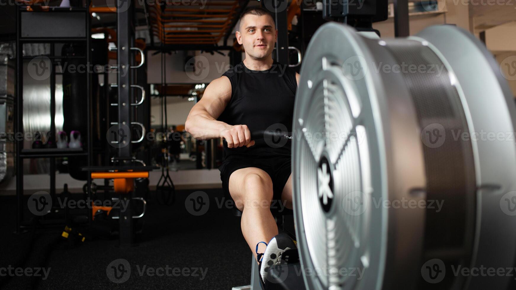 handsome male coach sits on a simulator in gym. Imitation rowing, boat with oars, for rowers. Smiling, happy, sports active healthy lifestyle, indoor concept, coaching and individual lessons. banner photo