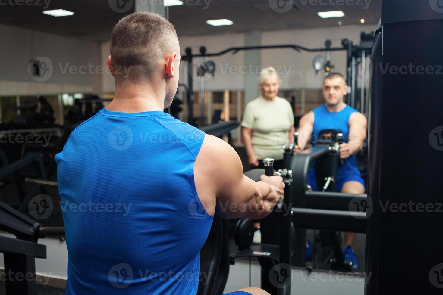 A handsome young coach man helps an old woman train sports in gym. Happy people smile, enjoy life. Active, healthy lifestyle, senior. rear view photo