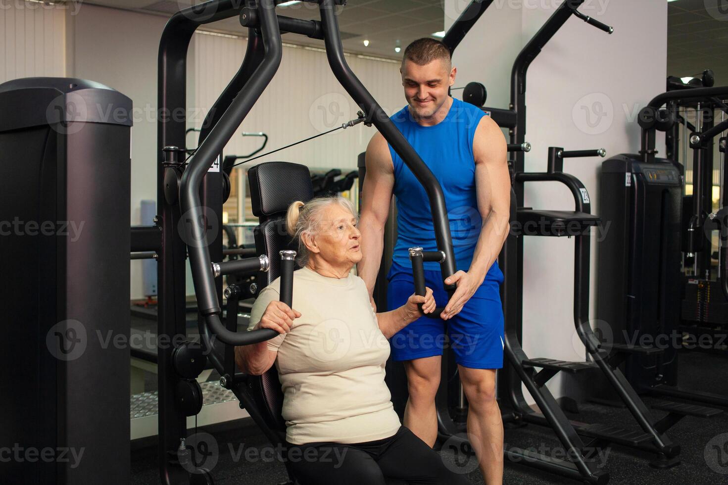 A handsome young coach man helps an old woman train sports in gym. Happy people smile, enjoy life. Active, healthy lifestyle, senior. photo