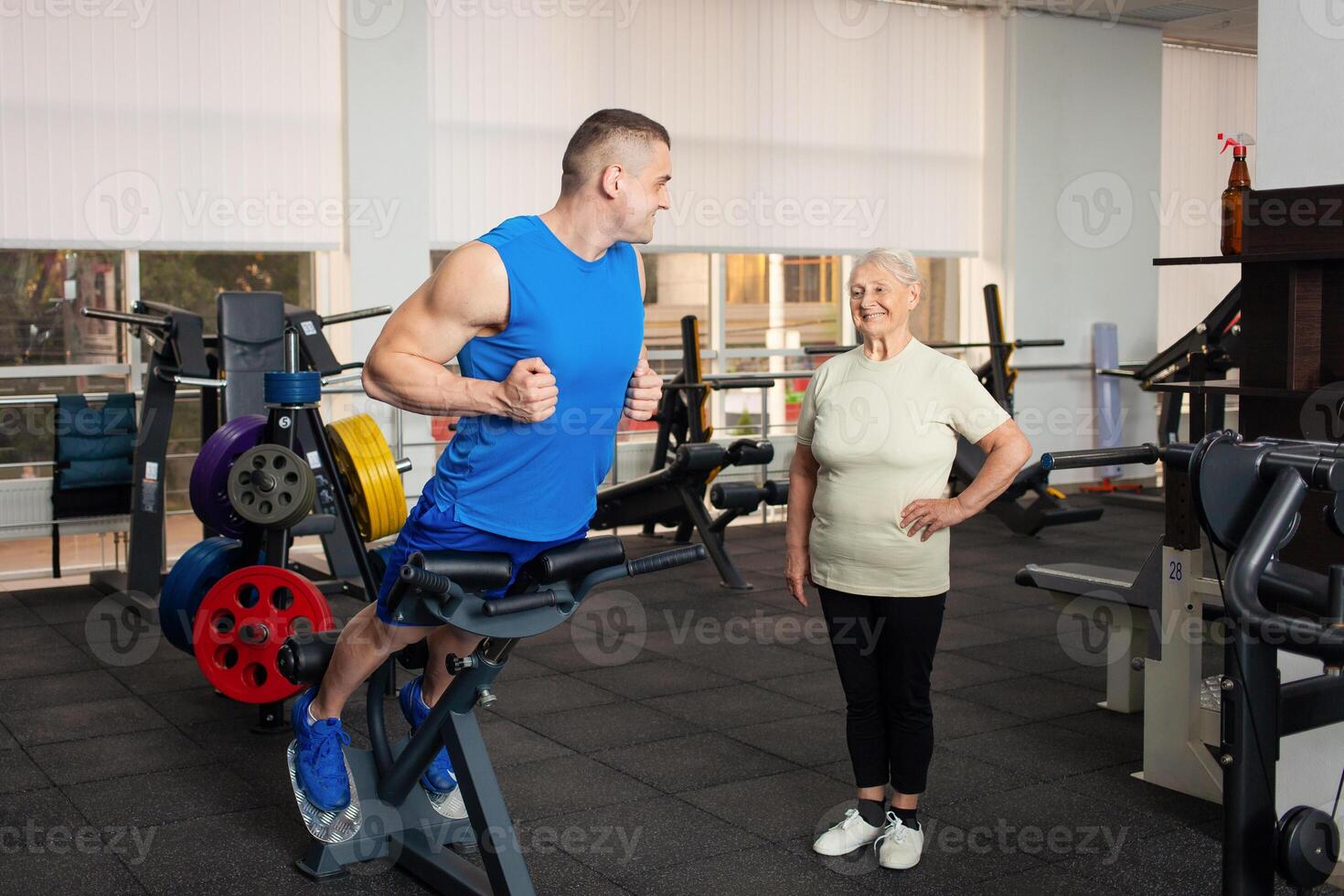 A beautiful young coach shows the old woman how to do the exercise on simulator. People smile, are happy, show class, thumb. Super. Sports active healthy lifestyle concept. photo