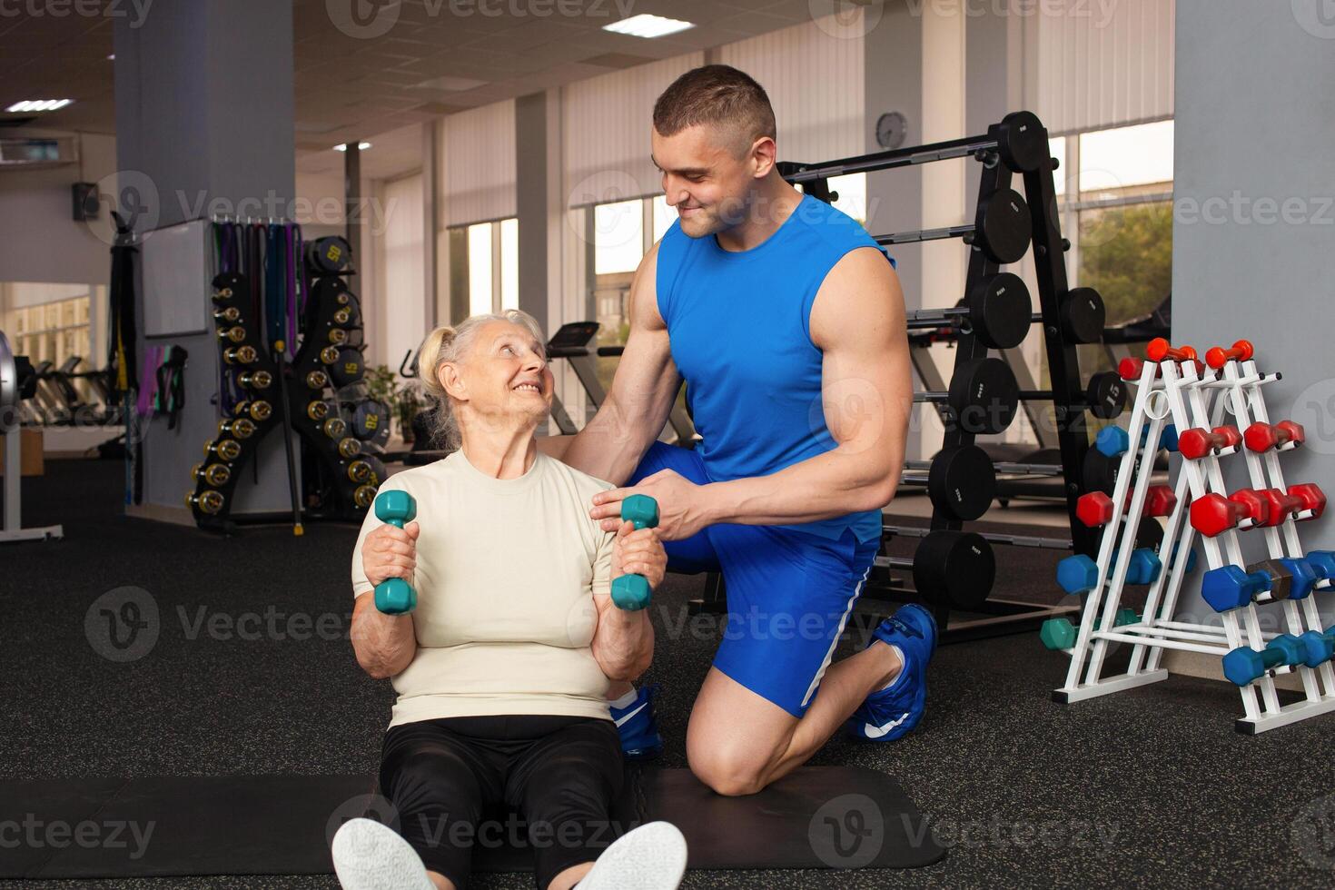 young male coach helps an old woman exercise in the gym. Active, sporty, healthy lifestyle. Senior concept. Indoor. Happy smiling people, retirement age. photo