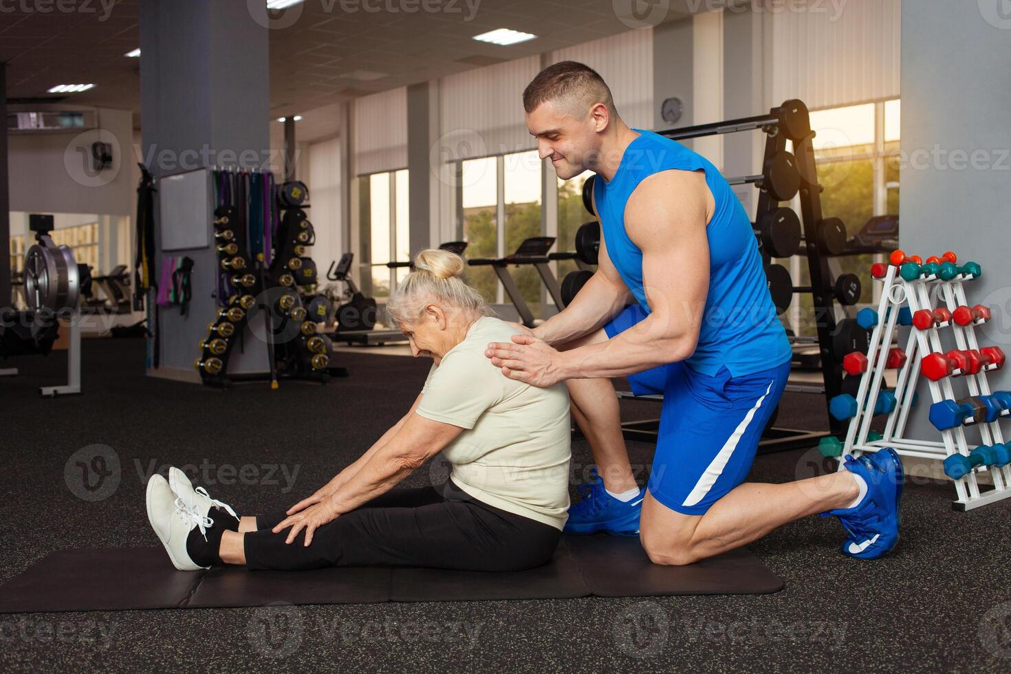 joven hermoso entrenador ayuda el más viejo mujer realizar extensión. aptitud gimnasia. gimnasia. activo, deportivo, sano estilo de vida en Jubilación edad. feliz, sonriente. físico educación para personas mayores en el estera foto