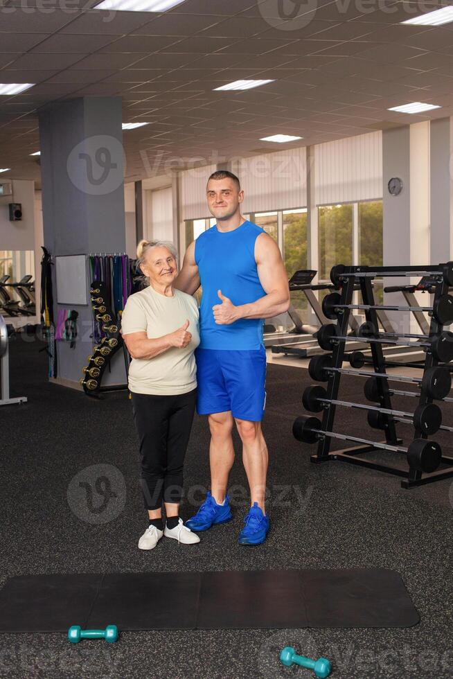 Portrait of a young coach in uniform and an old woman in gym. Physical culture for older people. Training and individual courses. Happy people smile, sport is cool, super, thumbs up. photo