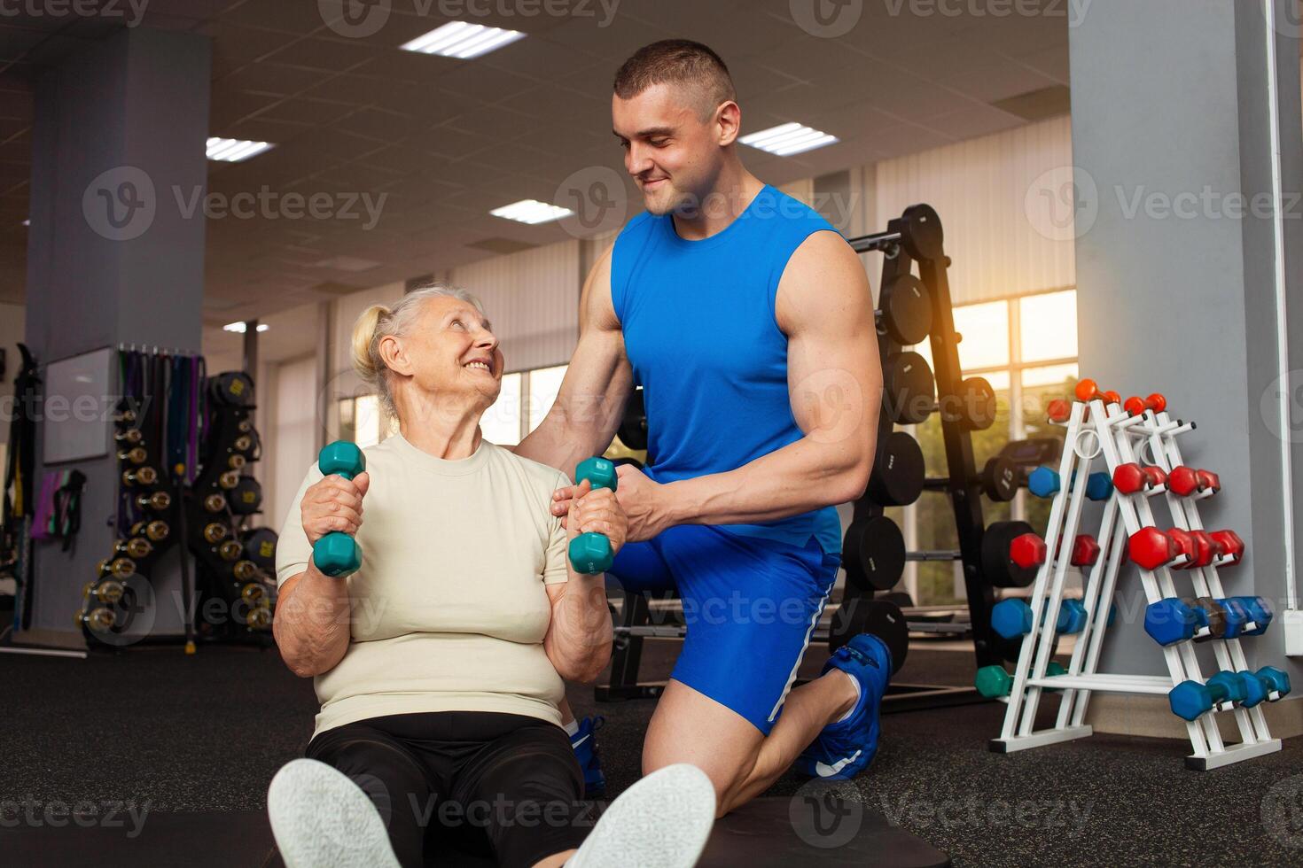 joven masculino entrenador ayuda un antiguo mujer ejercicio en el gimnasia. activo, deportivo, sano estilo de vida. mayor concepto. interior. contento sonriente gente, Jubilación edad. mancuernas, músculo columpio foto