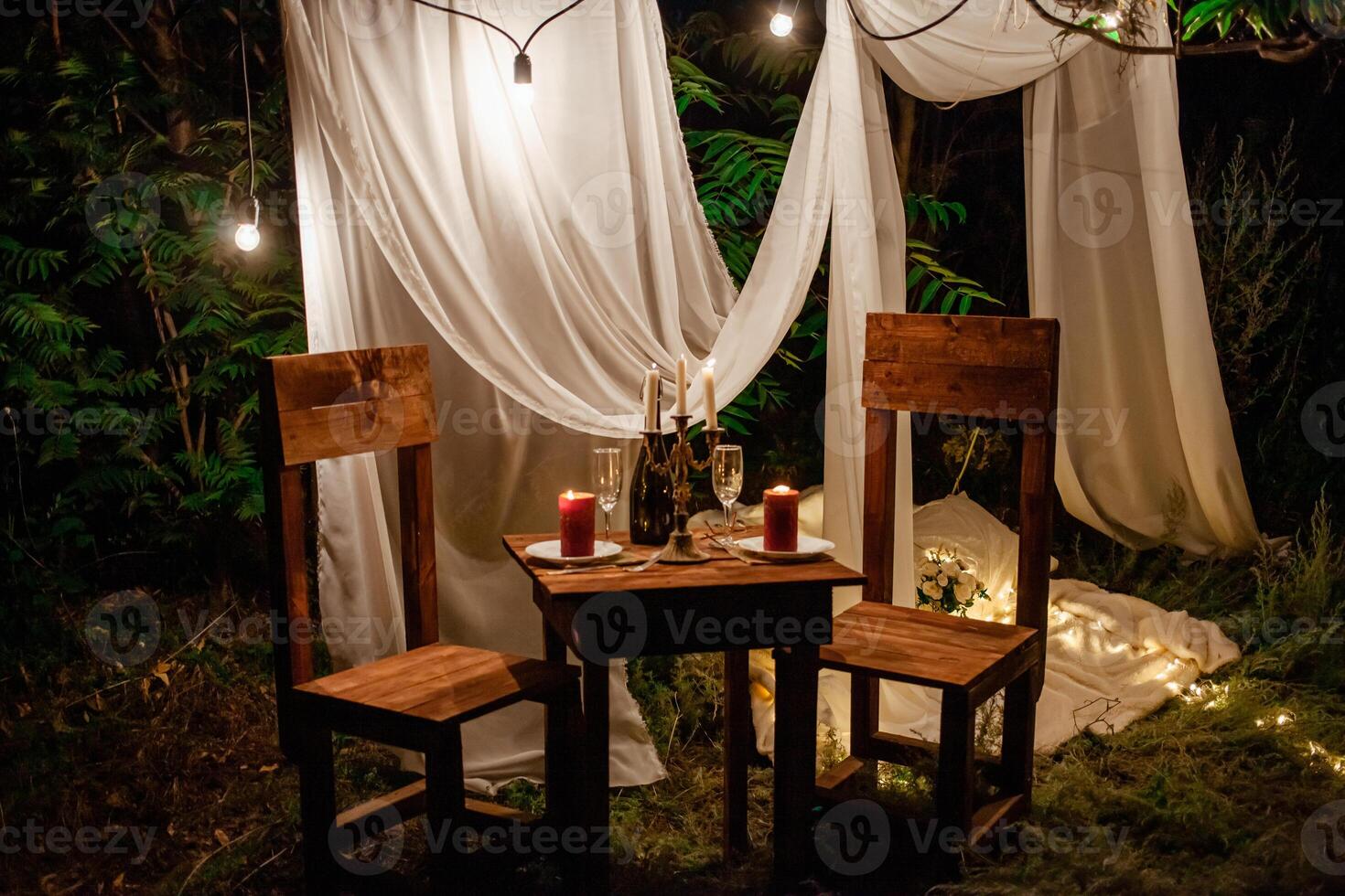 Table in the woods, romantic dinner for two by candlelight. White curtains on the tree, a garland of light bulbs, glasses, wine. Evening photo zone