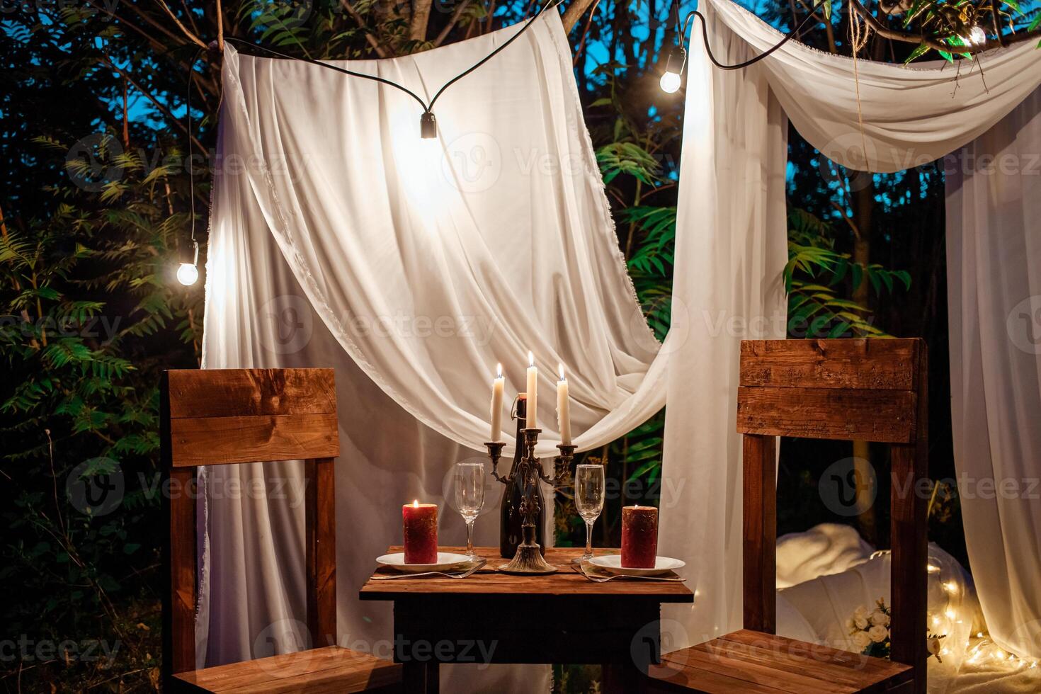 Table in the woods, romantic dinner for two by candlelight. White curtains on the tree, a garland of light bulbs, glasses, wine. Evening photo zone