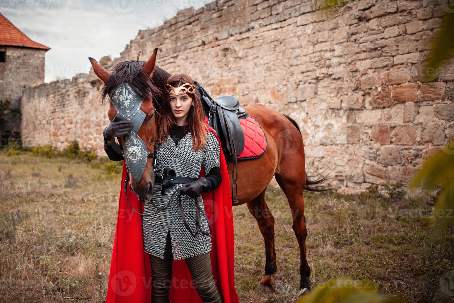 hermosa princesa con rojo capa soportes siguiente a el caballo en contra el fondo de un torre y un Roca pared foto