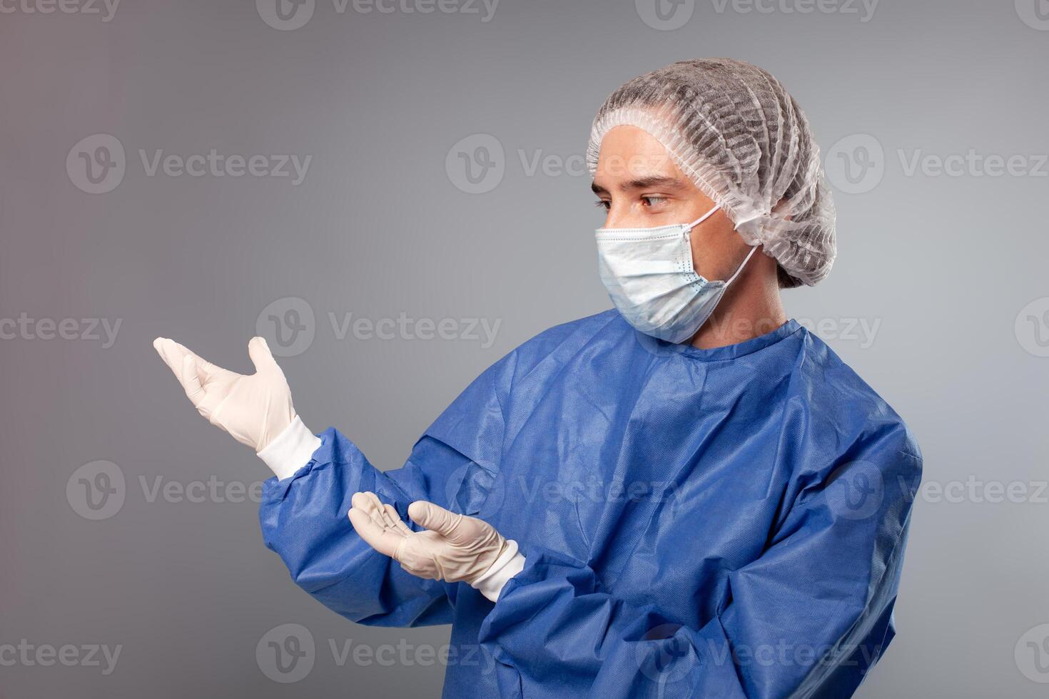 un hermoso masculino cirujano es un médico en un gris antecedentes en el estudio. un hombre en un túnica, un médico mascarilla, un sombrero. vacío espacio para texto. un hombre puntos su manos a alguna cosa a su bien. foto