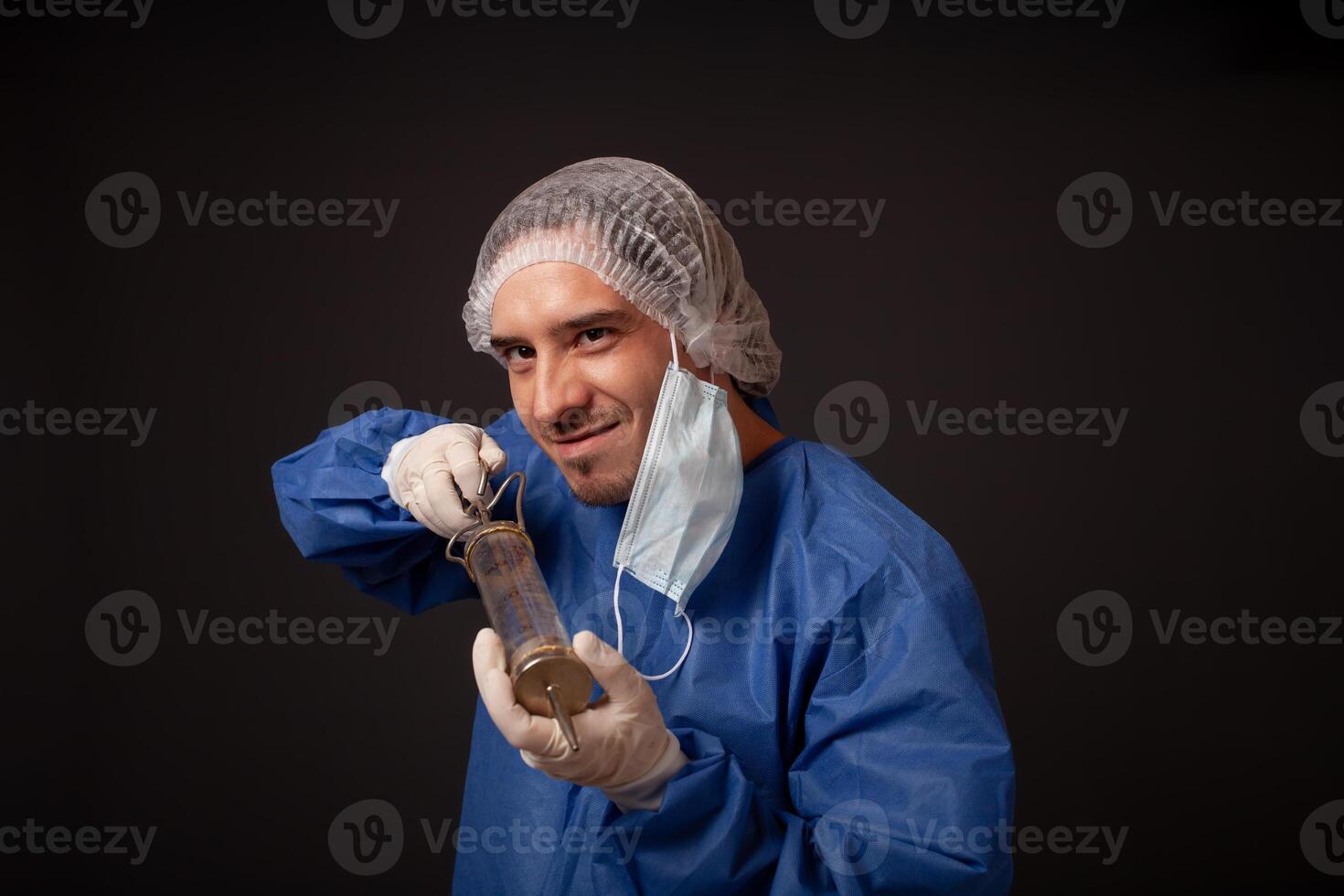 A male doctor with a terrible old syringe in his hands. The doctor in a medical mask, hat, robe and gloves. A terrible. The doctor scares with a huge syringe with a thick needle. photo