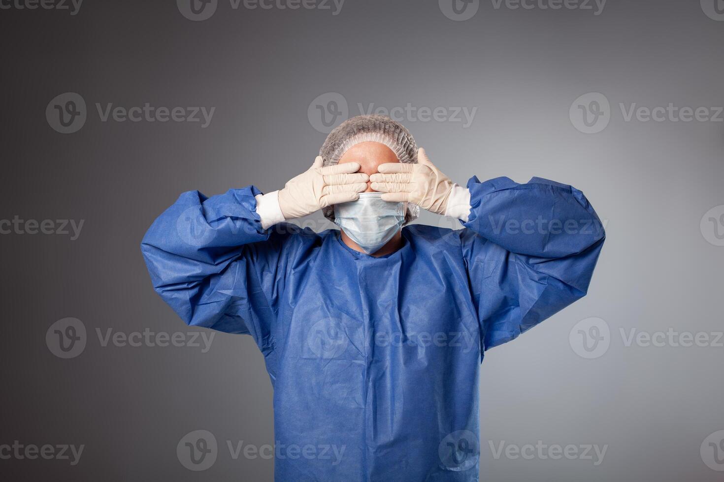 A handsome male surgeon is a doctor on a gray background in the studio. A man in a robe, a medical mask, a hat. Empty space for text. A man covers his eyes with his hands, does not want to see. photo