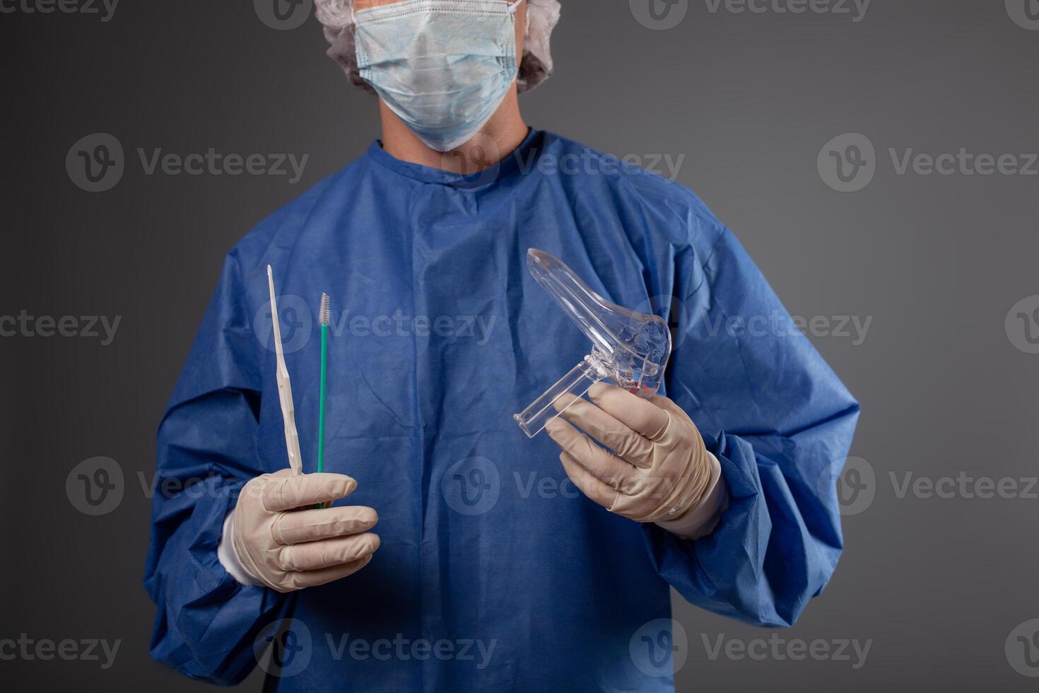 A gynecologist with a venus mirror in his hands. Gynecological accessories, brush, stick. A doctor in a medical mask, a robe, gloves, with a gynecological mirror on a gray background. photo