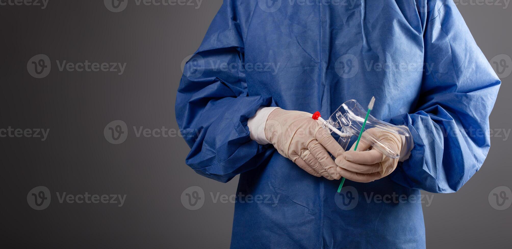 A gynecologist with a venus mirror in his hands. Gynecological accessories, brush, stick. A doctor in a medical mask, a robe, gloves, with a gynecological mirror on a gray background. photo
