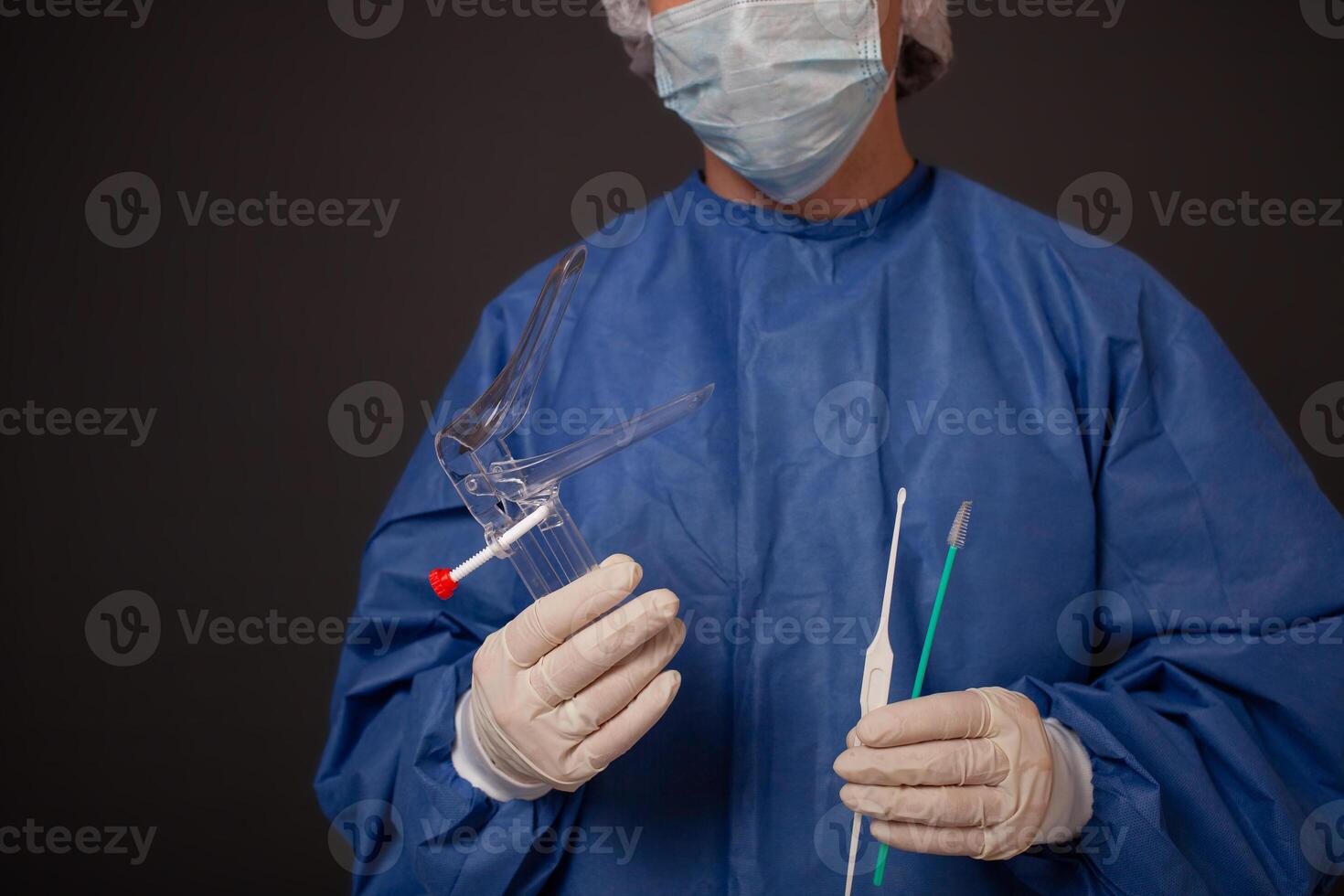 A gynecologist with a venus mirror in his hands. Gynecological accessories, brush, stick. A doctor in a medical mask, a robe, gloves, with a gynecological mirror on a gray background. photo