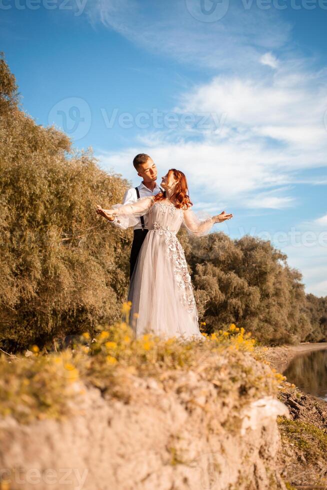 un joven Boda Pareja en amor en el borde de un acantilado en contra el fondo de el río y el cielo. un mujer en un hermosa gris vestido, un hombre en un traje en el apuntalar de el lago. foto