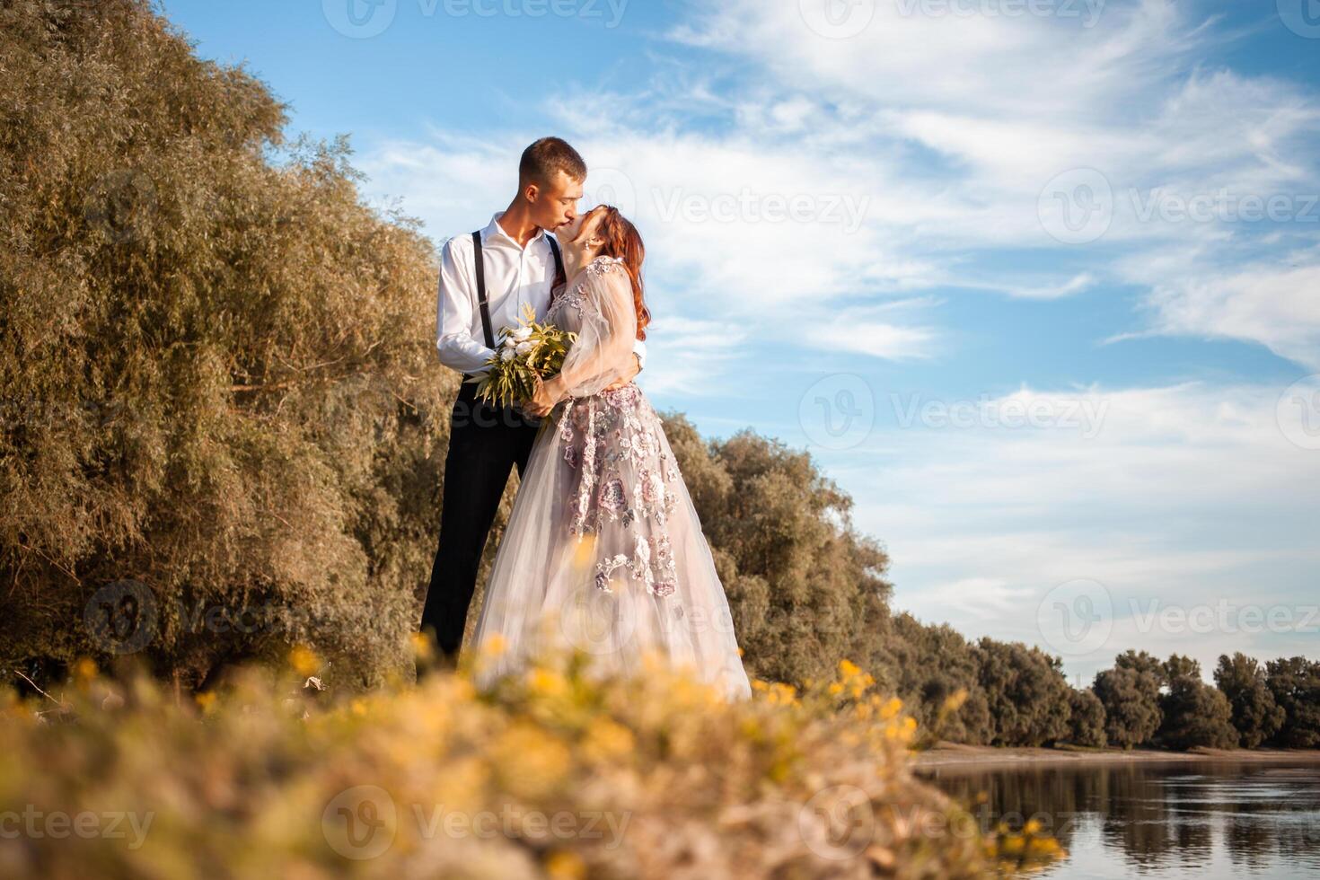 un joven Boda Pareja en amor en el borde de un acantilado en contra el fondo de el río y el cielo. un mujer en un hermosa gris vestido, un hombre en un traje en el apuntalar de el lago. foto