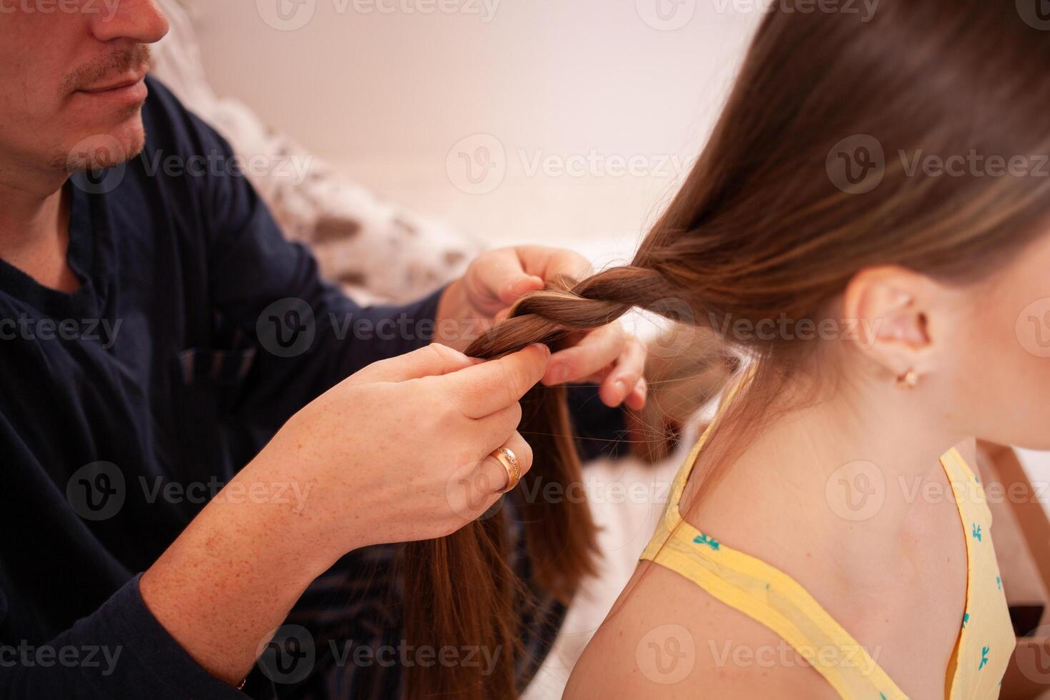 Dad in pajamas braids his daughter's pigtail in the morning. A man goes through his hair, does the hair of his daughter. photo