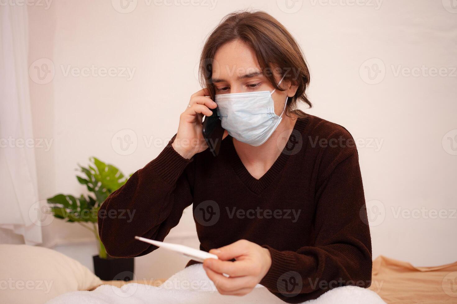 un enfermo hombre llamadas un ambulancia en el teléfono. un enmascarado hombre cheques el temperatura y diales el del doctor número en el teléfono. coronavirus, hogar cuarentena. foto