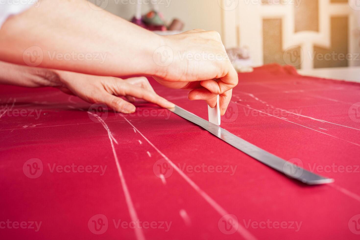 Seamstress at work. Dressmaker making clothes in modern studio. Tailor holding pencil and marking fabric. Woman standing at table with cut textile, sewing machine, thread, pins, needles, tape, cutouts photo