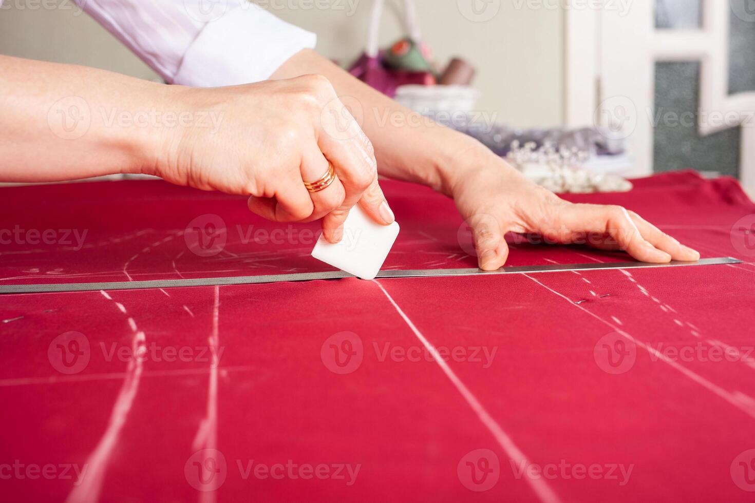 Seamstress at work. Dressmaker making clothes in modern studio. Tailor holding pencil and marking fabric. Woman standing at table with cut textile, sewing machine, thread, pins, needles, tape, cutouts photo