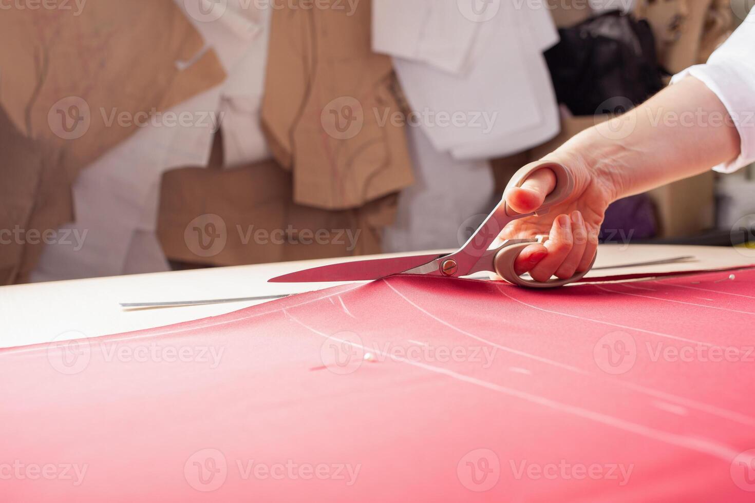 Seamstress at work. Dressmaker making clothes in modern studio. Tailor holding pencil and marking fabric. Woman standing at table with cut textile, sewing machine, thread, pins, needles, tape, cutouts photo