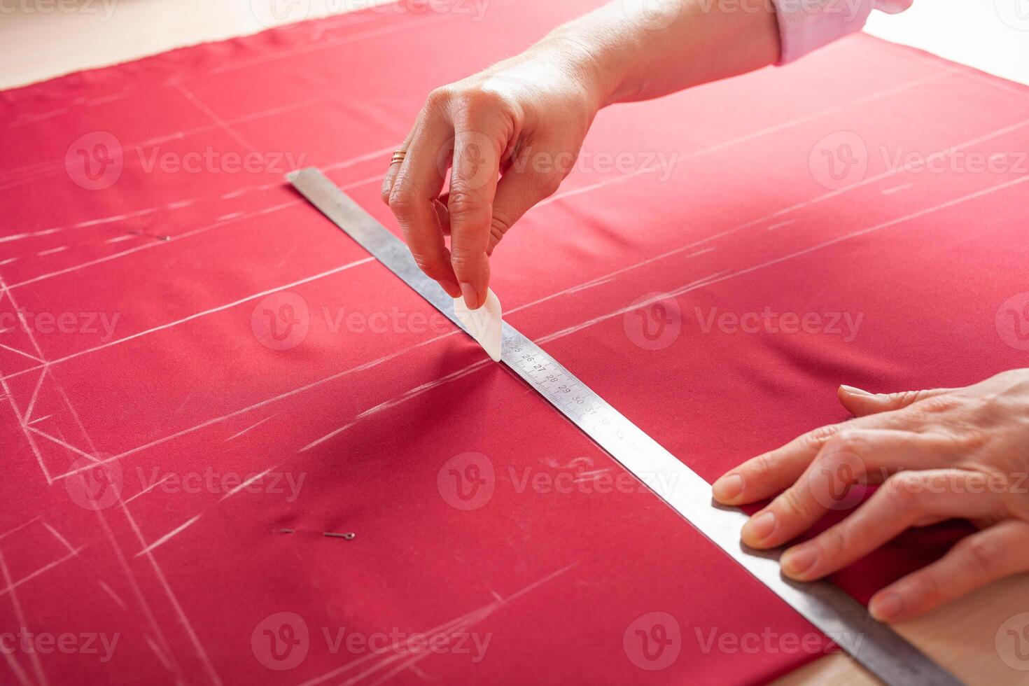 Seamstress at work. Dressmaker making clothes in modern studio. Tailor holding pencil and marking fabric. Woman standing at table with cut textile, sewing machine, thread, pins, needles, tape, cutouts photo