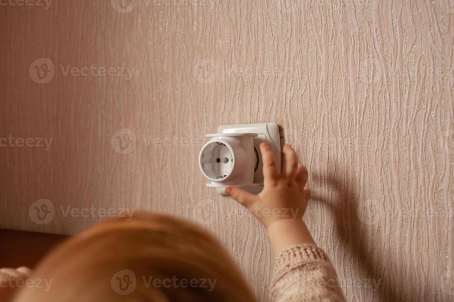 The child plays with an outlet, pulls his hands to it, sticks his fingers. The child is in danger at home. Baby fingers in the socket. photo