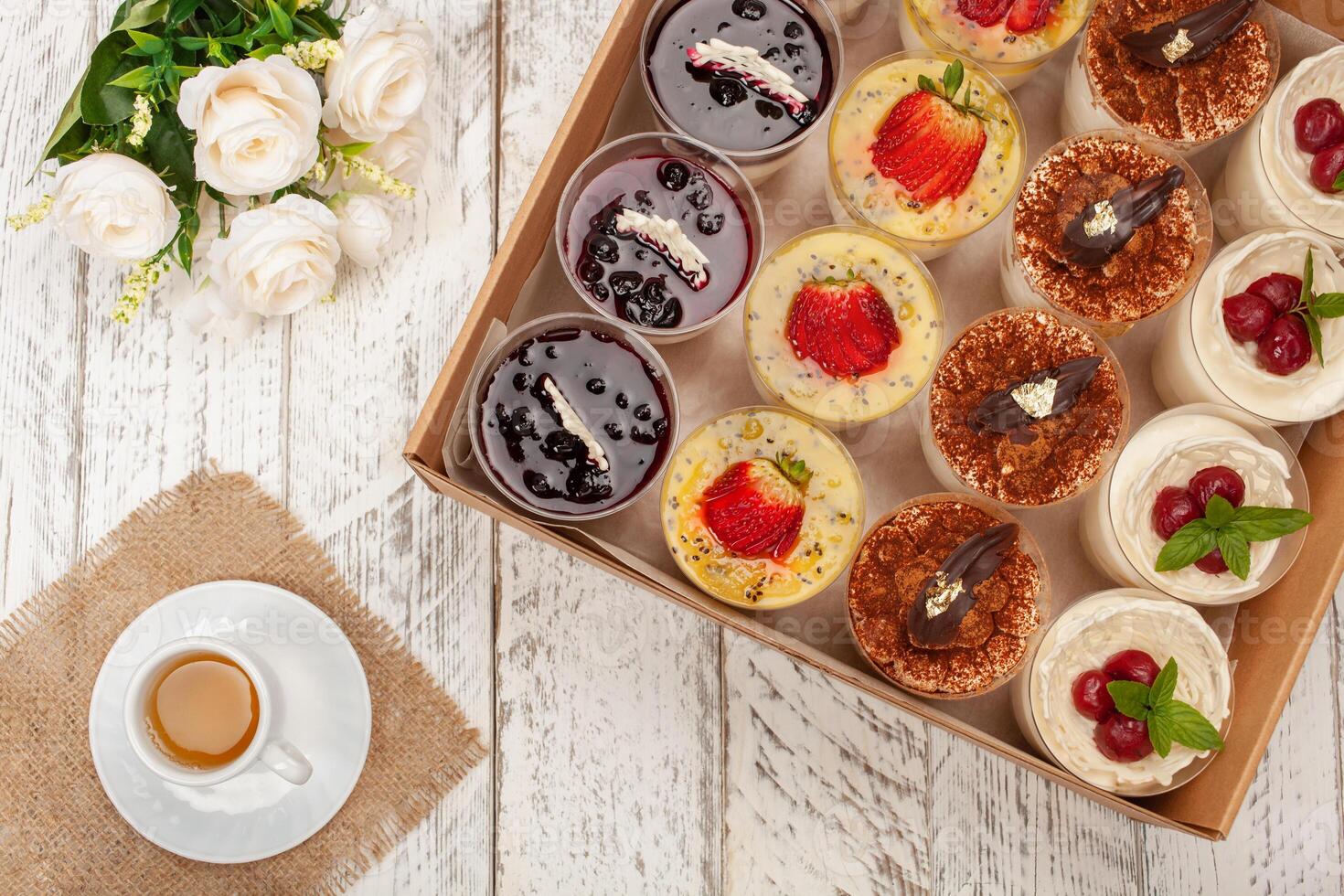 dulce postre en vaso con galleta, baya Fruta y azotado crema. un taza de té, flores, pasteles en un caja. entrega de comida a el casa para fiestas o un cena foto