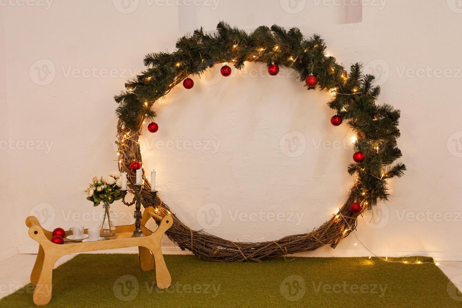 Christmas photo zone, lights, Christmas tree, huge New Year's wreath, tray with flowers, cups and candles. Green carpet, imitation of grass. Studio.