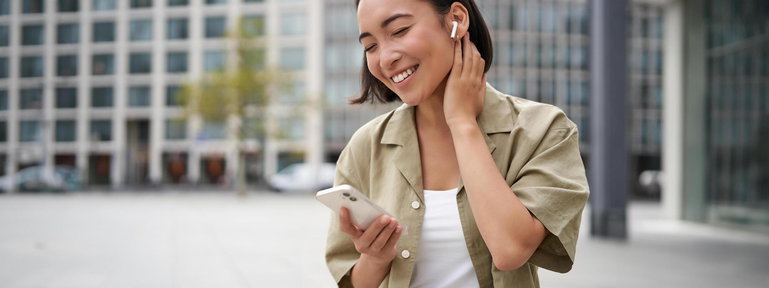 Smiling asian girl listens music in wireless headphones, looks at her phone, choosing music or podcast. Young woman calling someone, using headphones photo