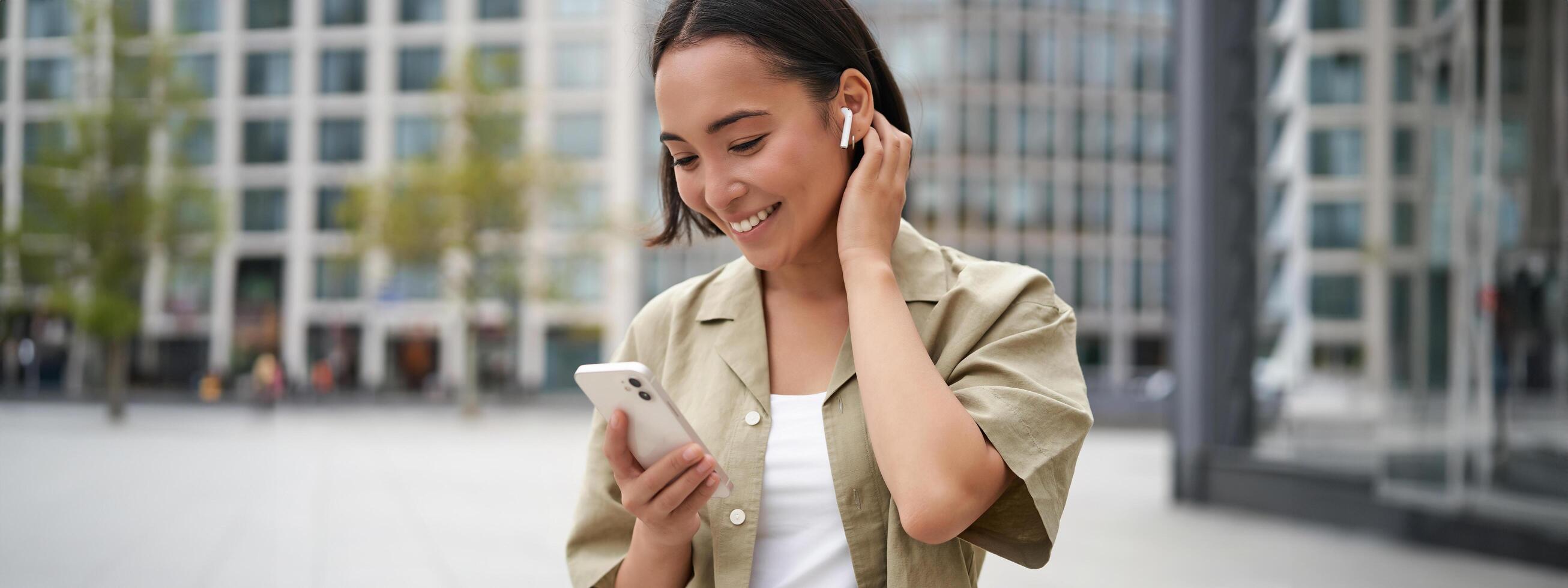 Smiling asian girl listens music in wireless headphones, looks at her phone, choosing music or podcast. Young woman calling someone, using headphones photo