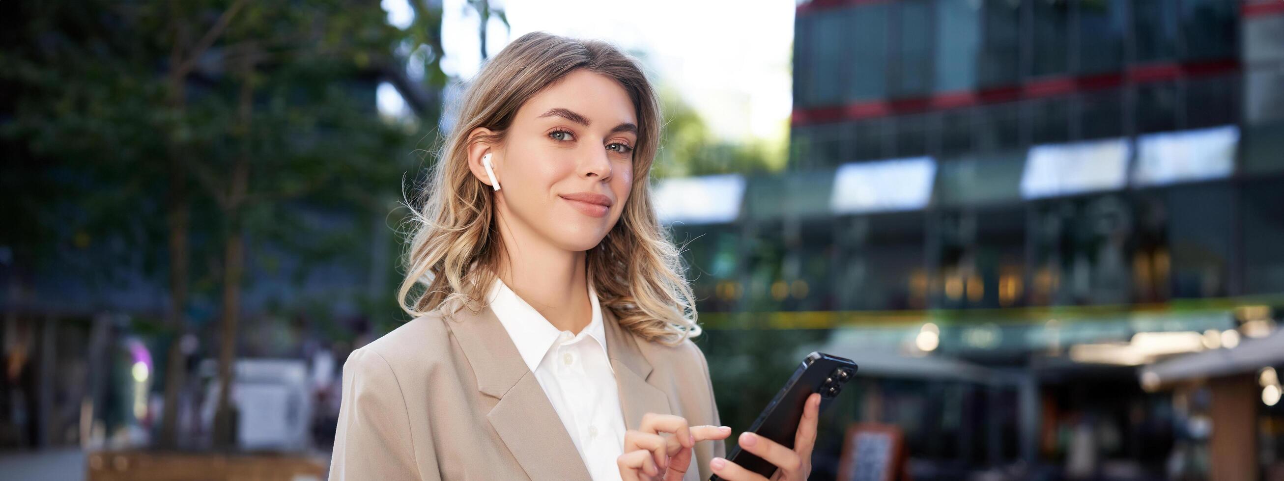 Smiling beautiful corporate woman in beige suit, standing on street in city with wireless headphones, holding smartphone photo