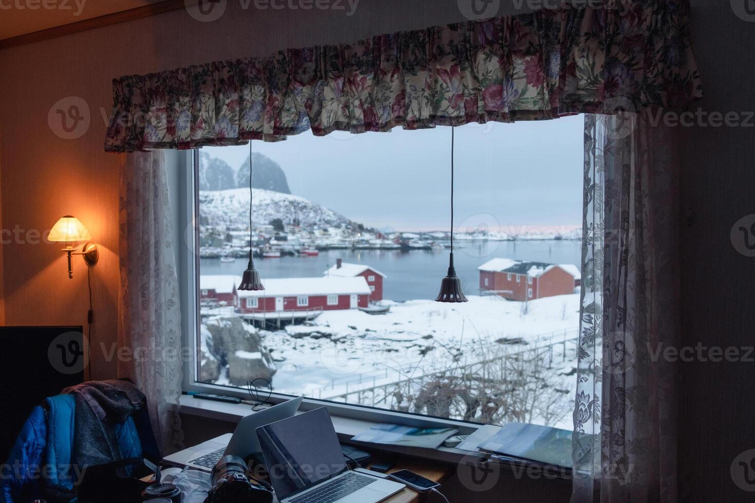 Interior of lamp shining with view fishing village on winter through window and laptop on desk in scandinavian house photo