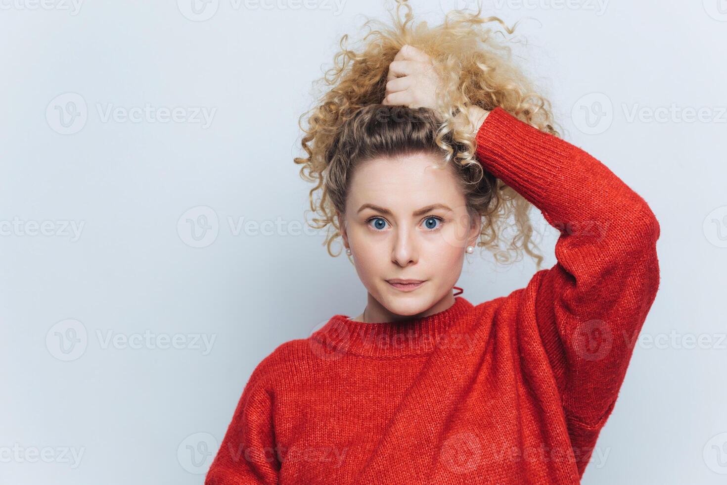 Curious woman with voluminous curly hair in a vibrant red sweater, hand in hair, looking thoughtfully aside photo