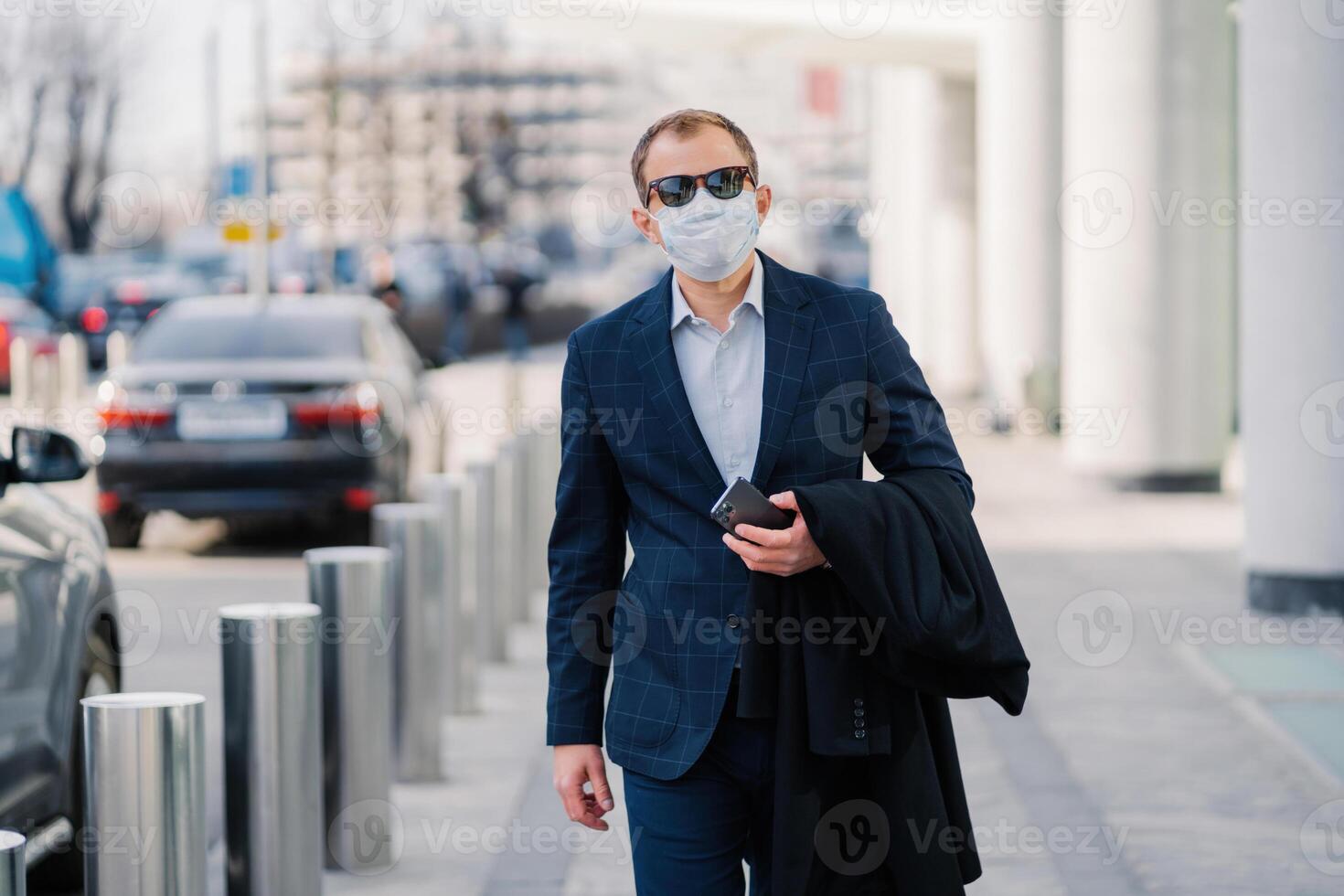 Professional man in a blue checkered suit walks confidently in the city, wearing a protective face mask and sunglasses, holding a phone. photo