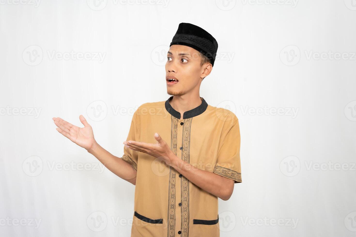 Asian Muslim man wearing brown Muslim clothes smiling happily while pointing at an empty space. Isolated white background. photo