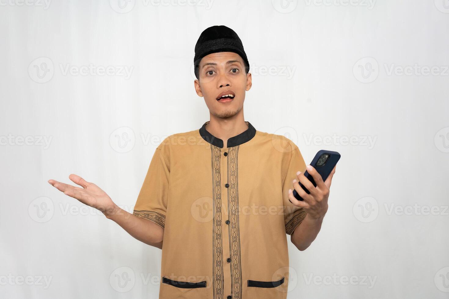 Asian Muslim man wearing Muslim clothes is shocked, confused and surprised while carrying a smartphone. Isolated white background. photo