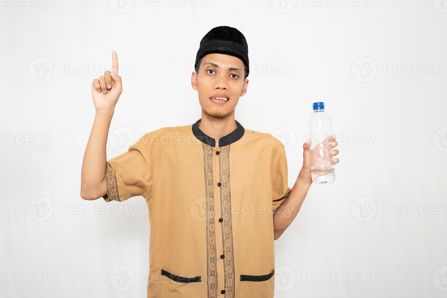 Asian Muslim man wearing Muslim clothes carrying mineral water in a bottle while pointing to an empty space. Isolated white background. photo