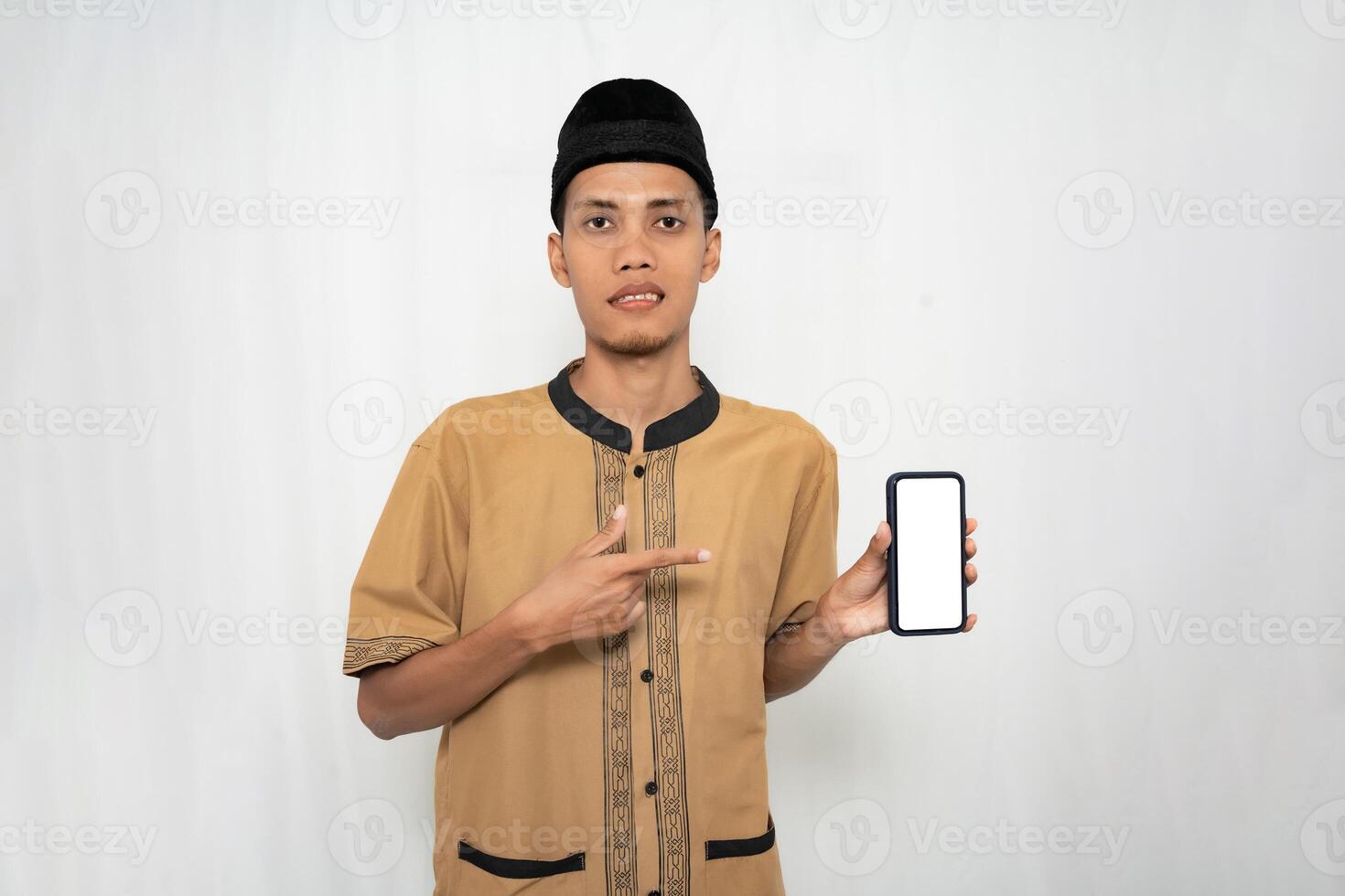 Asian Muslim man wearing brown Muslim clothes smiling happily while pointing at the smartphone screen. Isolated white background. photo