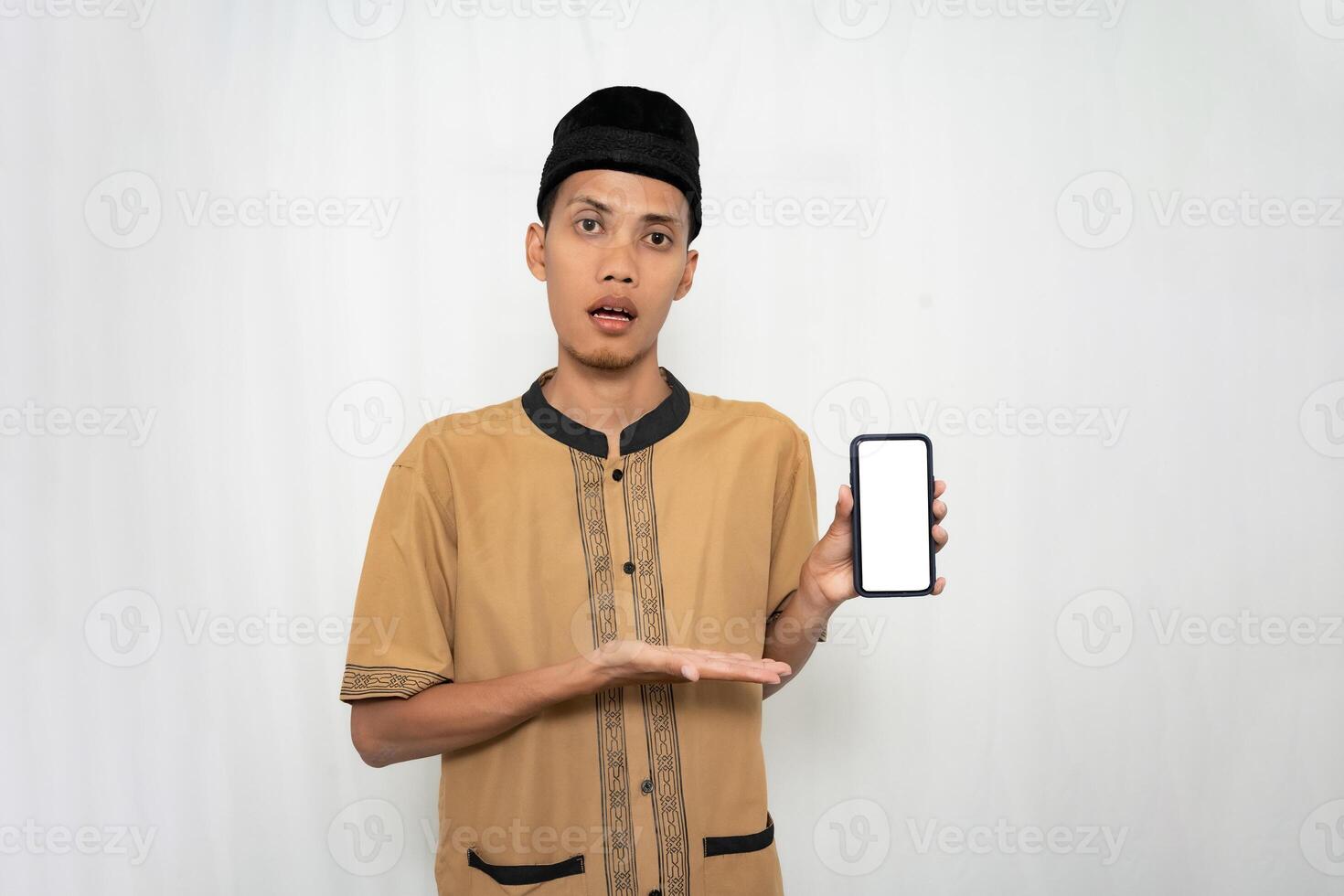 Asian Muslim man wearing brown Muslim clothes smiling happily while pointing at the smartphone screen. Isolated white background. photo