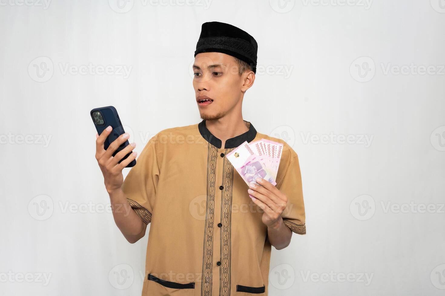 Asian Muslim man wearing Muslim clothes is shocked looking at the smartphone screen while carrying money in his other hand. Isolated white background. photo