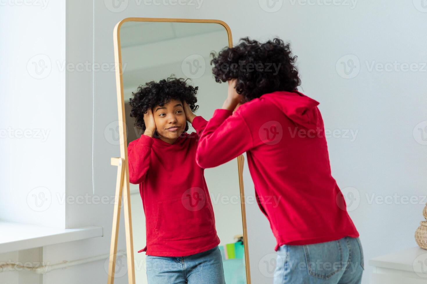 Love yourself. Beautiful young smiling african american woman dancing touching curly hair enjoying her mirror reflection. Black lady looking at mirror looking confident and happy. Self love concept photo