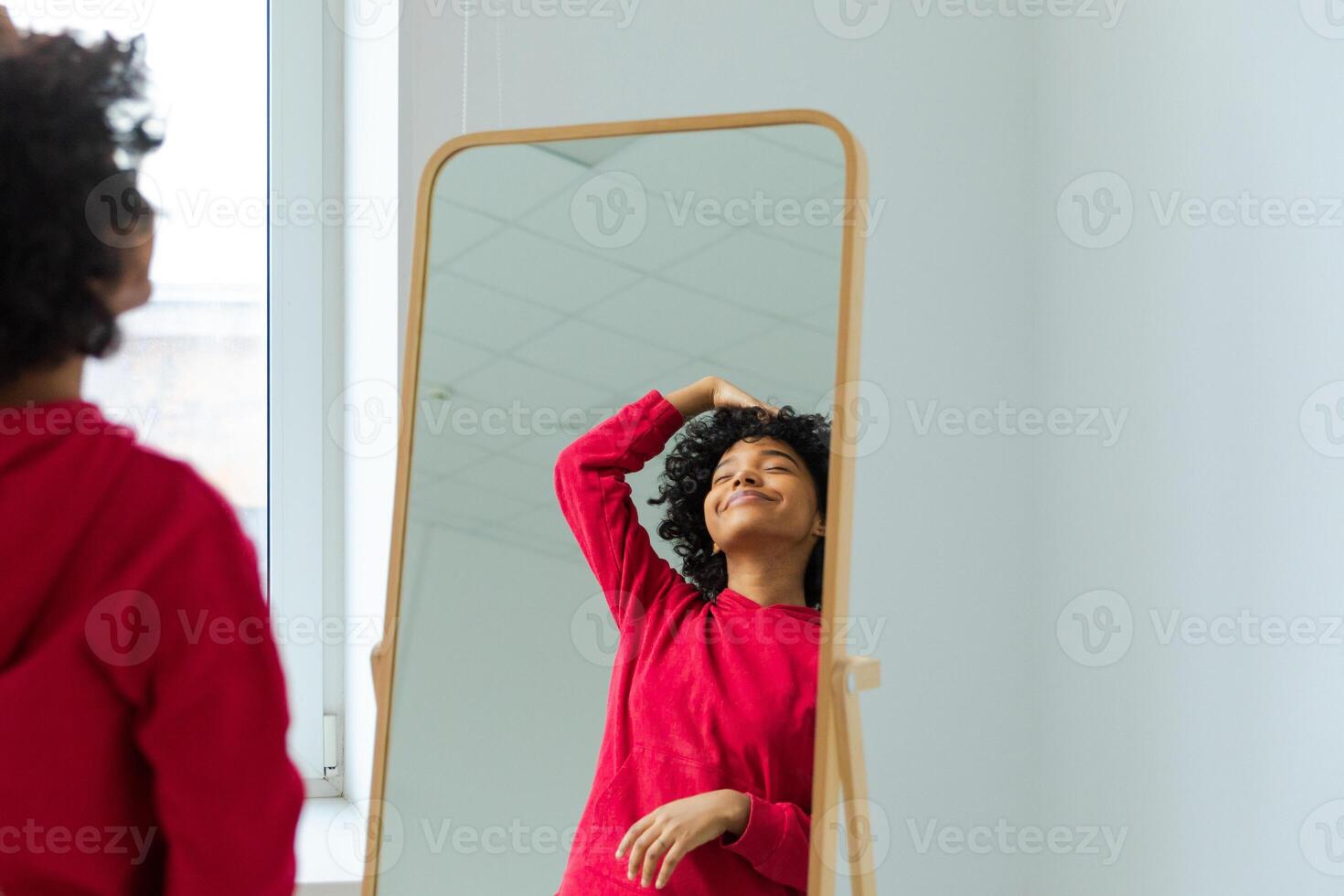 Love yourself. Beautiful young smiling african american woman dancing touching curly hair enjoying her mirror reflection. Black lady looking at mirror looking confident and happy. Self love concept photo