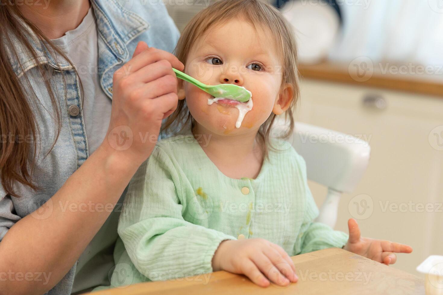Happy family at home. Mother feeding her baby girl from spoon in kitchen. Little toddler child with messy funny face eats healthy food at home. Young woman mom giving food to kid daughter photo