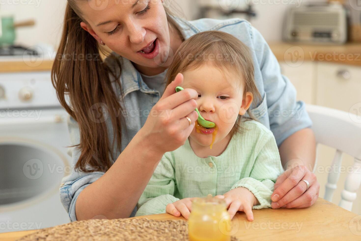 Happy family at home. Mother feeding her baby girl from spoon in kitchen. Little toddler child with messy funny face eats healthy food at home. Young woman mom giving food to kid daughter photo