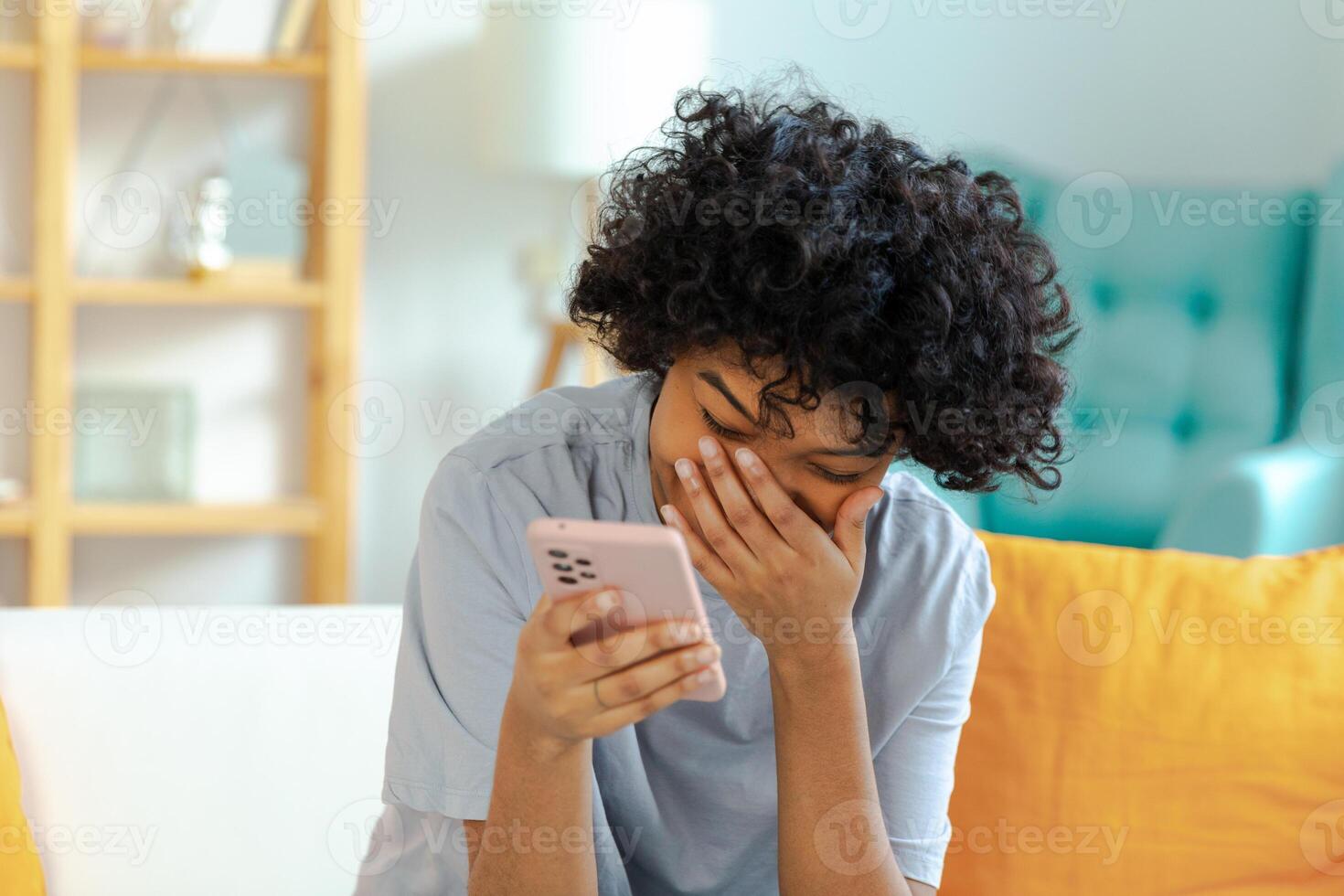 Excited happy young black african american woman holding cell phone laughing feeling joy getting mobile message. Overjoyed girl laughing aloud sitting on couch watching funny video reading news photo