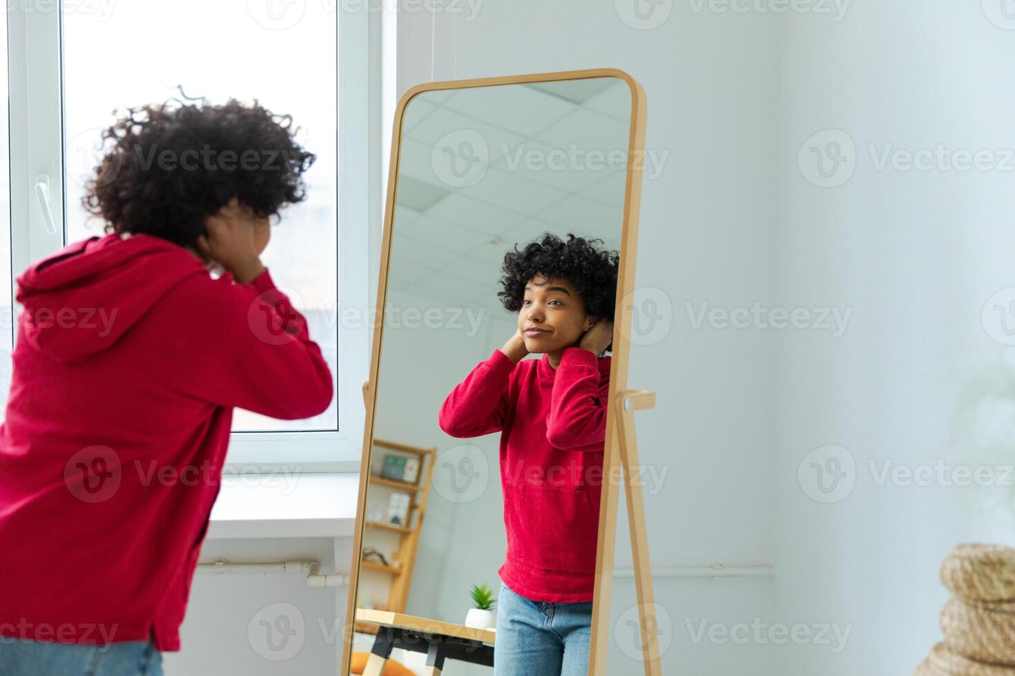 Love yourself. Beautiful young smiling african american woman dancing touching curly hair enjoying her mirror reflection. Black lady looking at mirror looking confident and happy. Self love concept photo